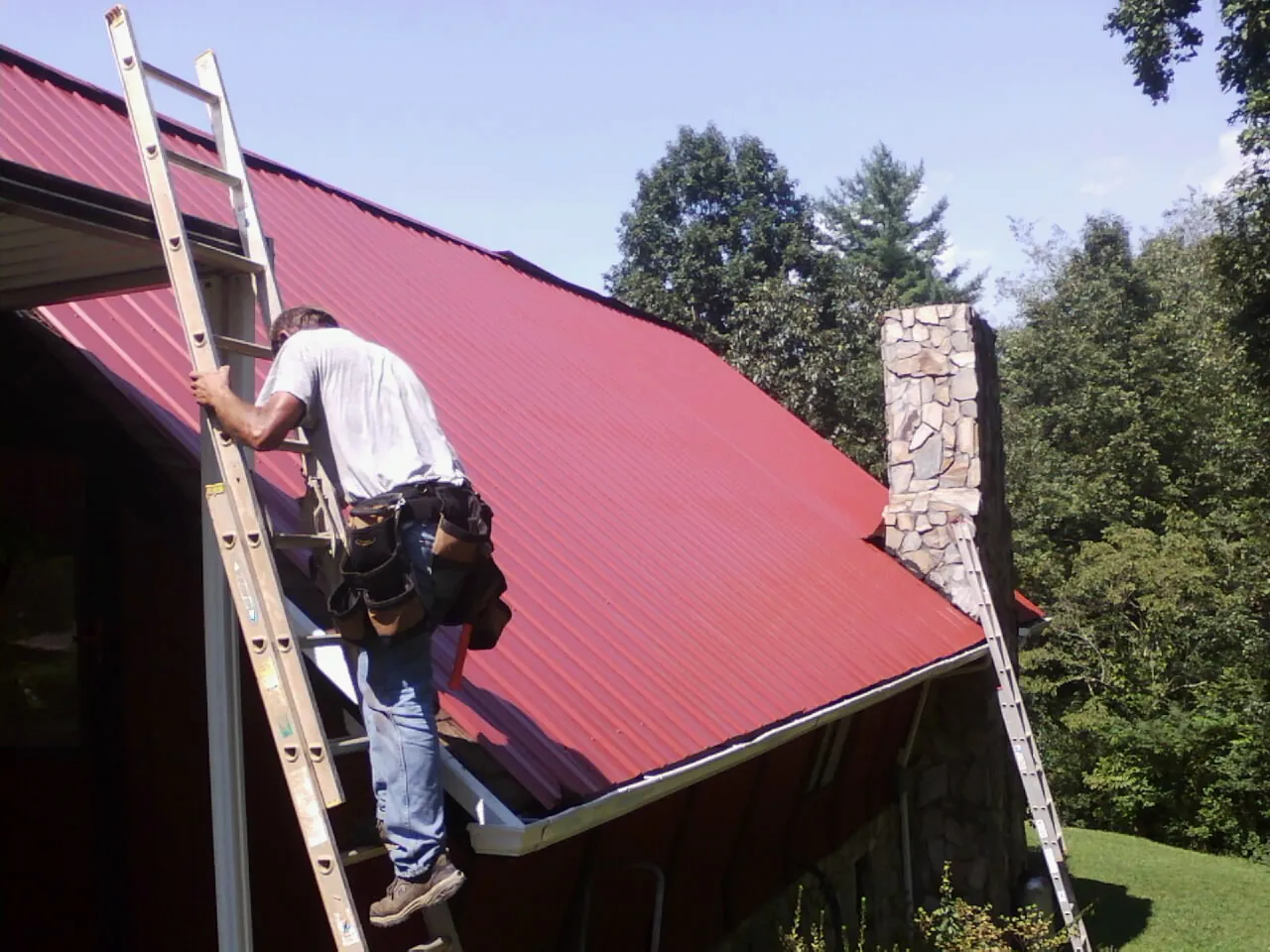 Roofer on a ladder beside a red metal roof and stone chimney — Soffit & Fascia Repair by Roofing Brothers in Bostwick